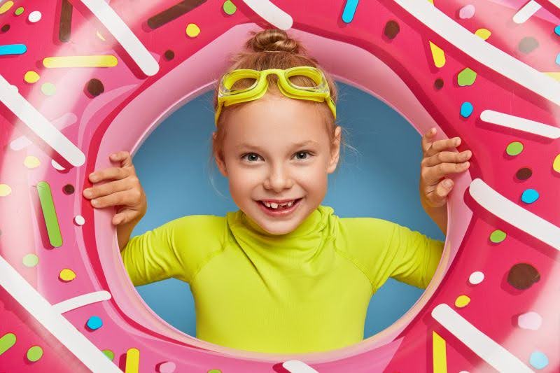 dievča s plaveckými okuliarmi sa pozerá cez nafukovacie koleso v tvare ružového donutu